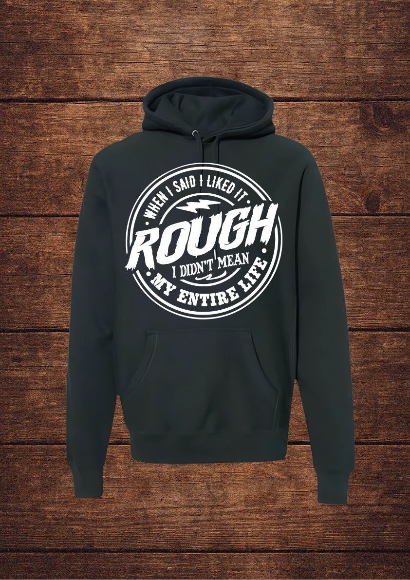 Black hoodie with black circular design and bold typography stating 'WHEN I SAID I LIKED IT ROUGH I DIDN'T MEAN MY ENTIRE LIFE'; features a drawstring hood and front pouch pocket, laid flat on rustic wood