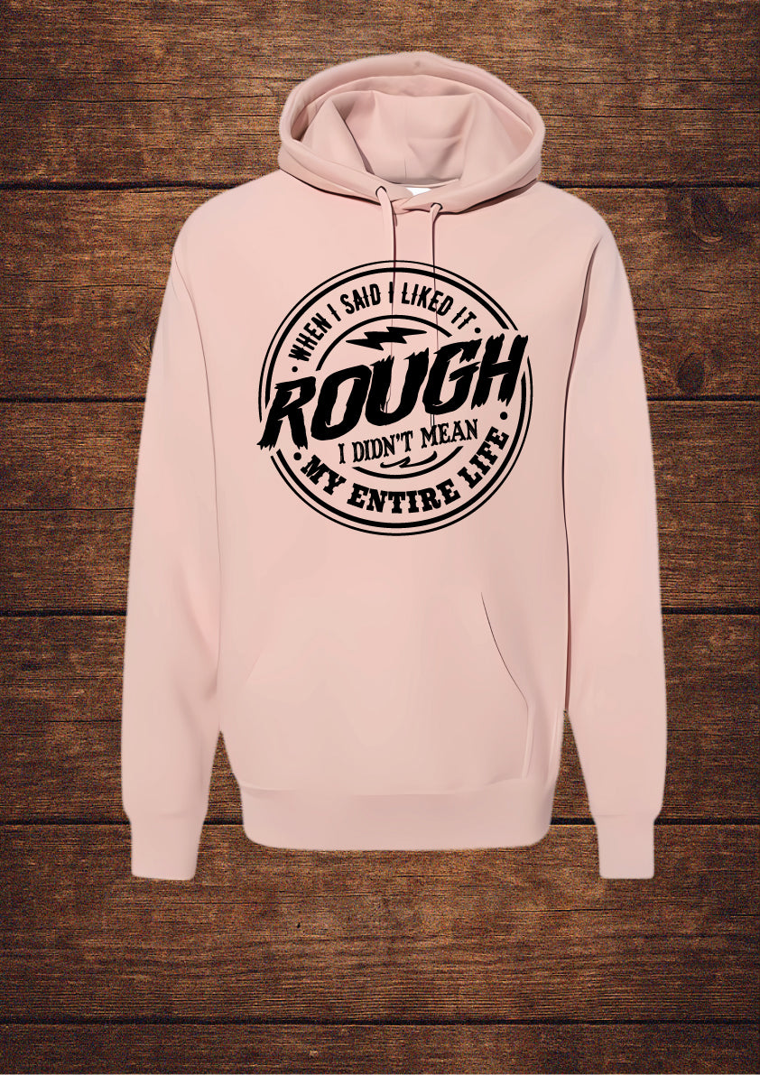Light Pink hoodie with black circular design and bold typography stating 'WHEN I SAID I LIKED IT ROUGH I DIDN'T MEAN MY ENTIRE LIFE'; features a drawstring hood and front pouch pocket, laid flat on rustic wood
