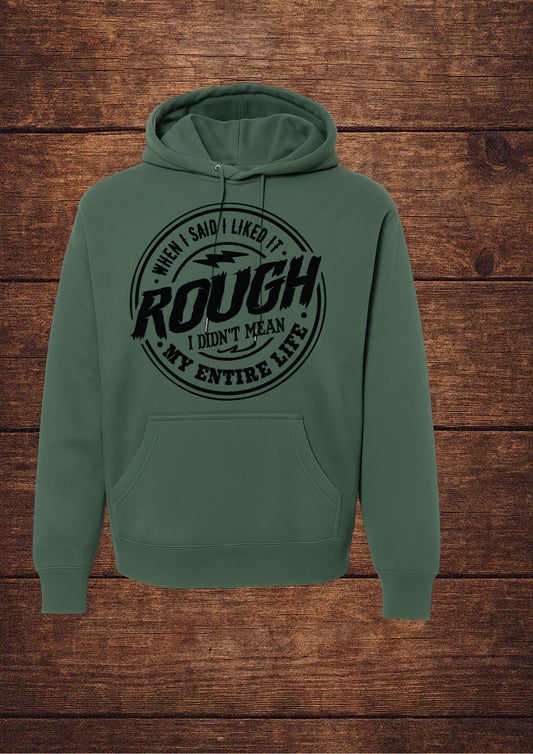 Green hoodie with black circular design and bold typography stating 'WHEN I SAID I LIKED IT ROUGH I DIDN'T MEAN MY ENTIRE LIFE'; features a drawstring hood and front pouch pocket, laid flat on rustic wood