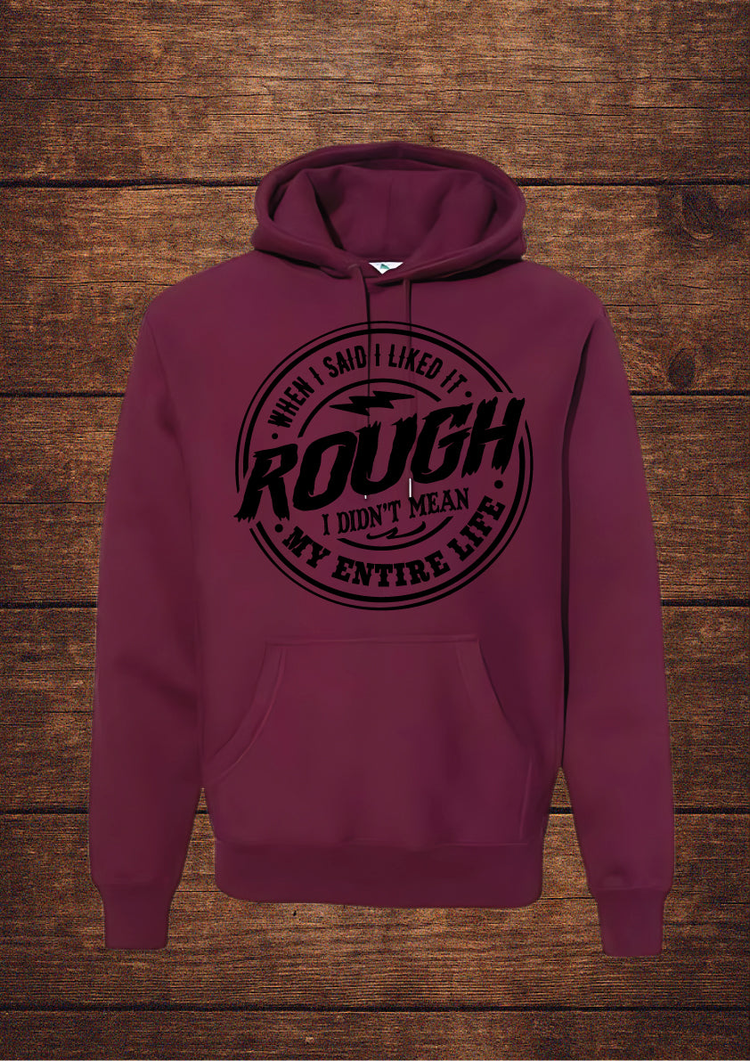 Maroon hoodie with black circular design and bold typography stating 'WHEN I SAID I LIKED IT ROUGH I DIDN'T MEAN MY ENTIRE LIFE'; features a drawstring hood and front pouch pocket, laid flat on rustic wood