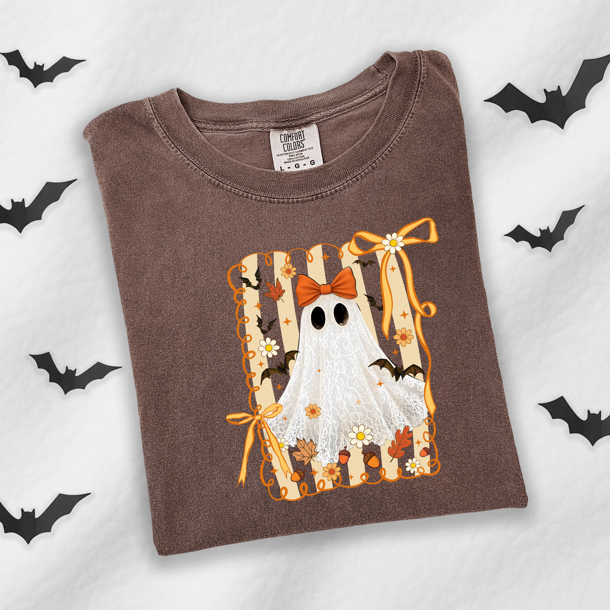 A brown crewneck t-shirt featuring a Halloween graphic of a white ghost with black eyes, an orange bow, and bats, laid flat on a white background with black bat silhouettes.