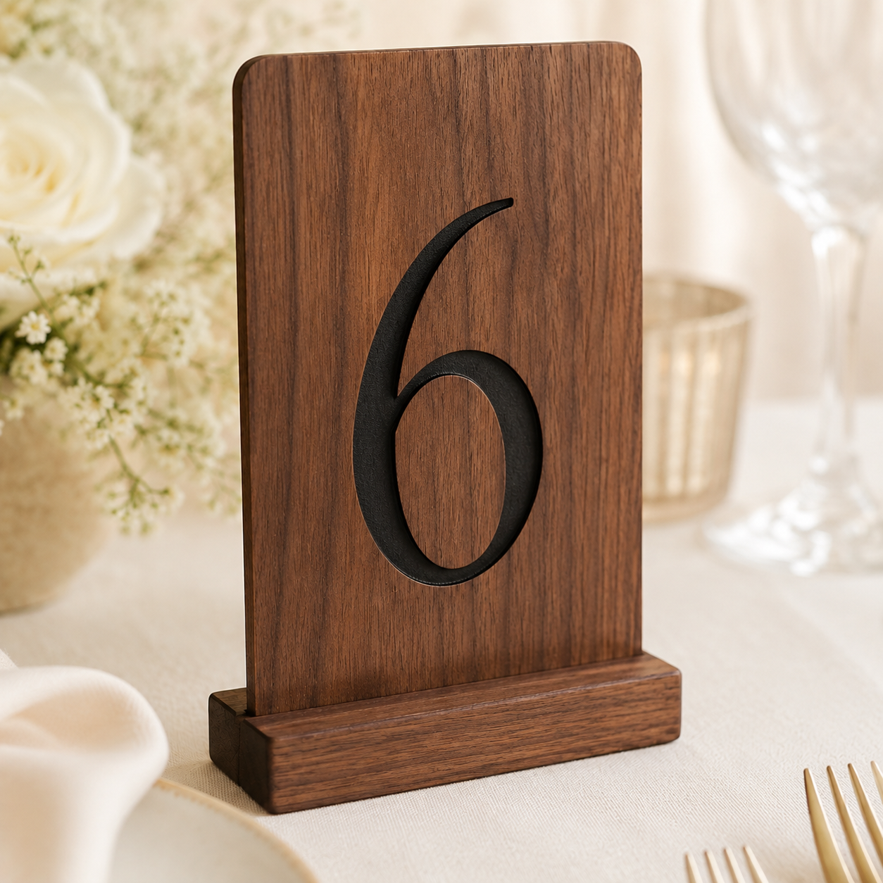 A wooden table number 6 stand with a dark brown wood grain finish and a black number cutout, displayed on a beige tablecloth with blurred floral and glassware in the background.
