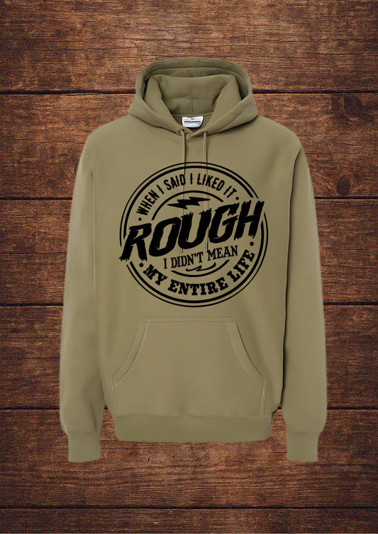 Olive hoodie with black circular design and bold typography stating 'WHEN I SAID I LIKED IT ROUGH I DIDN'T MEAN MY ENTIRE LIFE'; features a drawstring hood and front pouch pocket, laid flat on rustic wood