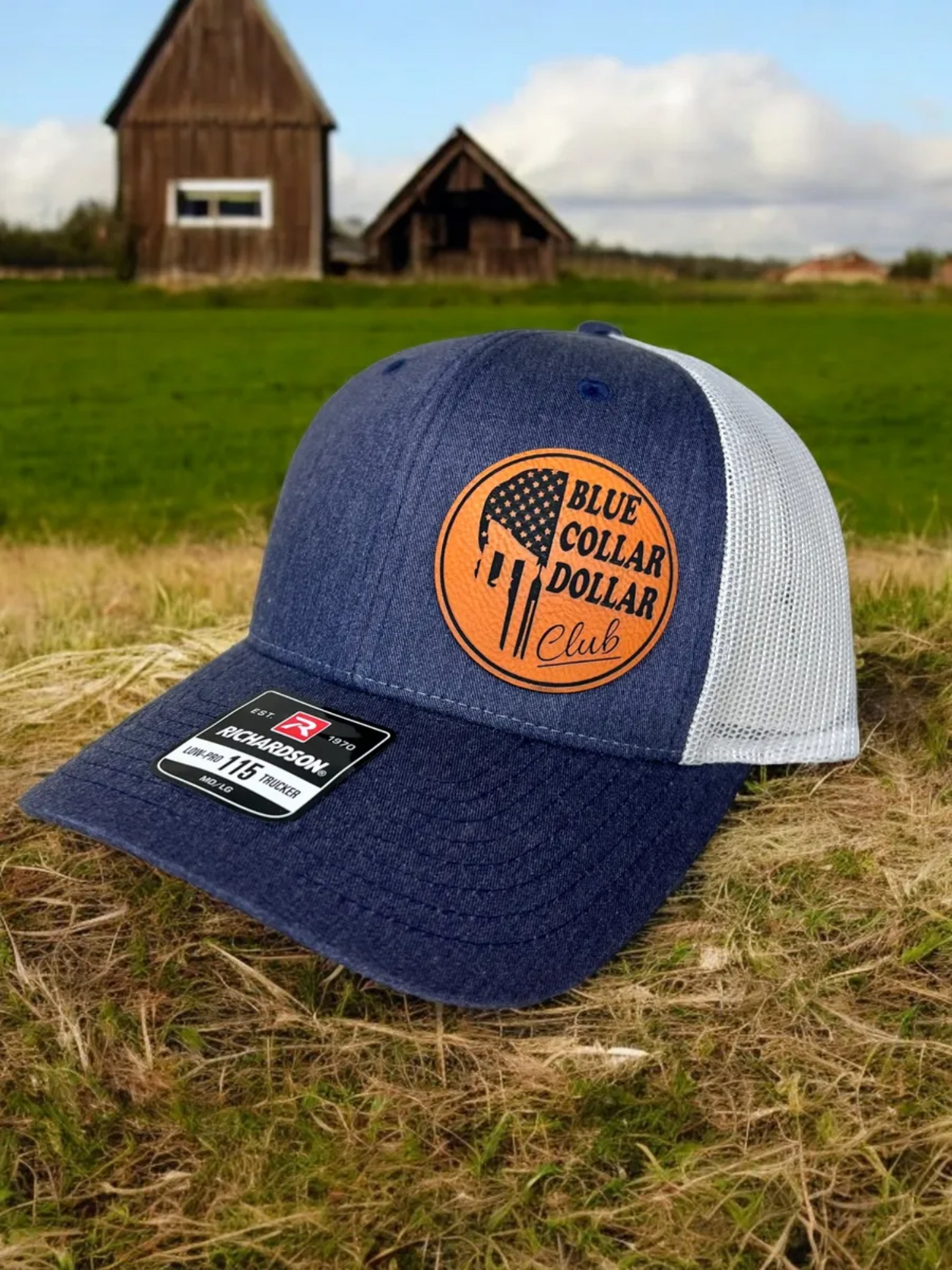 Blue and white cap with 'Blue Collar Dollar Club' logo on grass with barns in the background