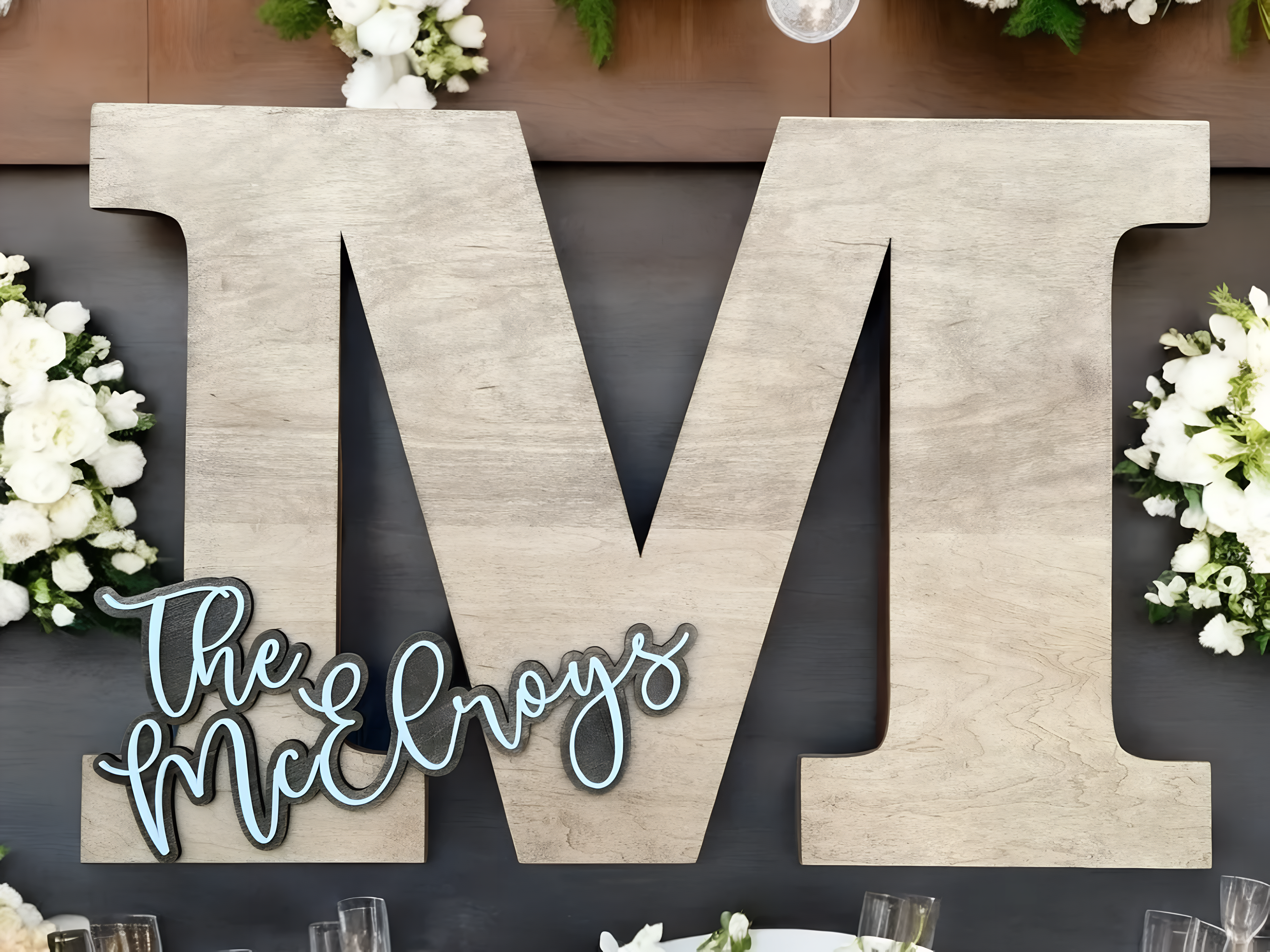 Decorative wooden letters spelling 'M' with 'The McElroys' on a dark background with floral arrangements.